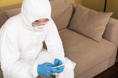 Lastik eldivenli adam, tıbbi maske ve tehlikeli madde giysisi. Evdeki kanepede akıllı telefon kullanıyor. 
