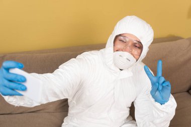 Tehlikeli madde giysisi ve tıbbi maske takmış bir adamın seçmeli odak noktası. Kanepede akıllı telefonuyla selfie çekerken barış işareti gösteriyor. 