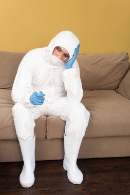 Tehlikeli madde giysisi ve tıbbi maskeli üzgün adam evdeki kanepede termometre tutuyor. 