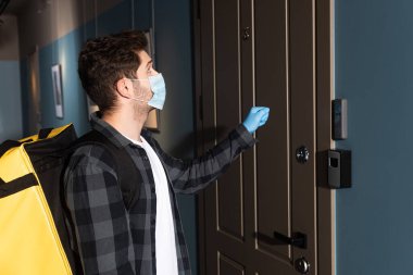 Tıbbi maskeli teslimatçı ve kapısında termal torba olan lateks eldivenli adamın yan görüntüsü.