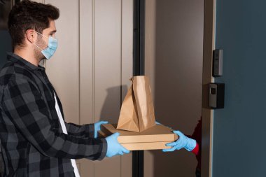 Tıbbi maskeli kuryenin açık kapının yanındaki kadına paket ve pizza kutuları verirken yan görünümü