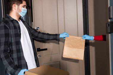 Teslimatçı adamın açık kapının yanındaki lateks eldivenli kadına paketi verirken yan görünüm 