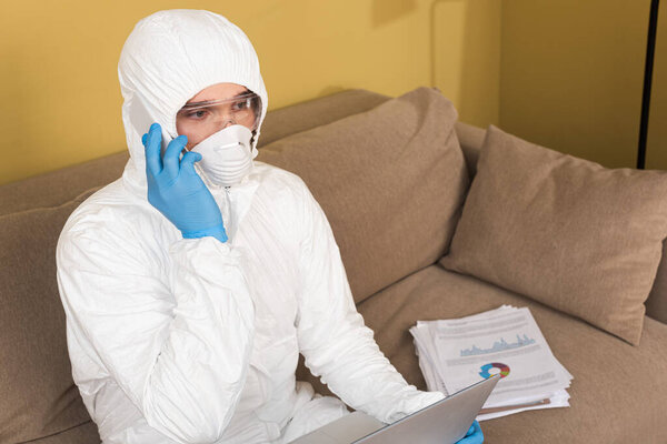 Freelancer in safety goggles, medical mask and hazmat suit talking on smartphone near laptop and documents on couch 