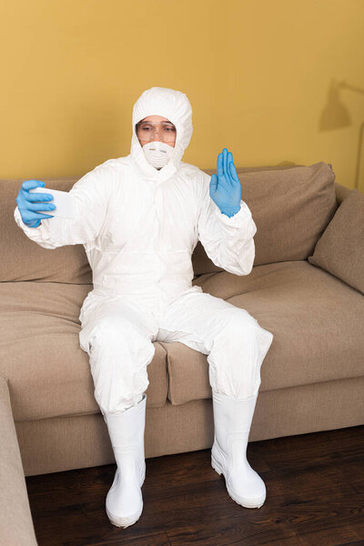 Man in hazmat suit and medical mask waving hand while taking selfie with smartphone on couch at home 