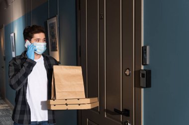 Tıbbi maskeli kurye girişte paket ve pizza kutularını tutarken akıllı telefondan konuşuyor.