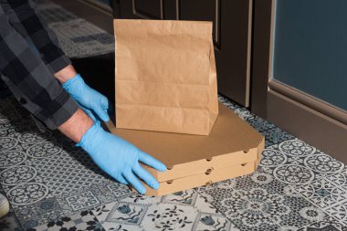 Lastik eldivenli teslimatçı pizza kutularını ve paketi kapının yakınına koyuyor. 