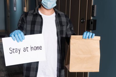 Tıbbi maskeli kuryenin elinde paket ve kartla evde kalıp girişte yazı yazması.