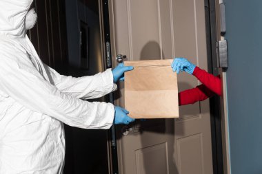 Tehlikeli madde giysisi ve tıbbi maskeli bir adamın açık kapının yanındaki lateks eldivenli bir kadına paket verişi. 