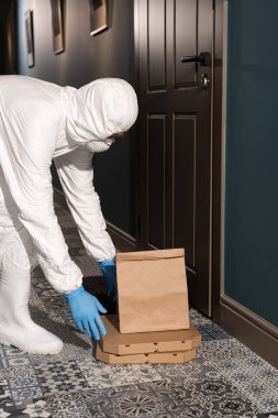 Tehlikeli madde giysisi ve sağlık maskesi takmış teslimatçının verandanın yanındaki yere paket ve pizza kutuları koyuşunun yan görüntüsü.