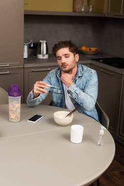 Hastalıklı adam boynuna dokunuyor ve termometreyi akıllı telefon ve mısır gevreğinin yanında tutuyor. 