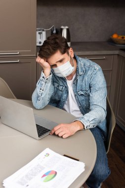 Medikal maskeli üzgün tele-işçi mutfak masasında çizelgeleri olan kağıtların yanındaki dizüstü bilgisayara bakıyor. 