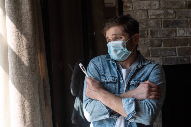 Maskeli hasta adam oturma odasının penceresinin yanında çapraz kollu termometre tutuyor.