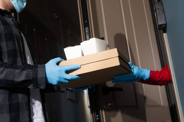 Обрезанный вид курьера в медицинской маске, дающего коробки пиццы и пакеты женщине в латексных перчатках возле открытой двери
