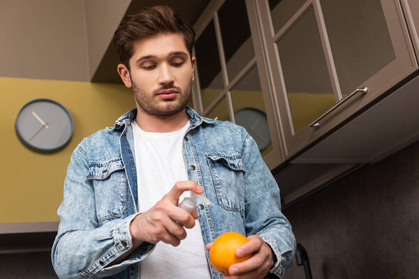Low angle view of handsome man using antiseptic and holding orange in kitchen 