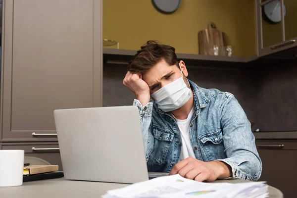 戴着医疗面具的悲伤自由职业者带着文件和笔记本电脑坐在桌旁的选择性焦点 — 图库照片