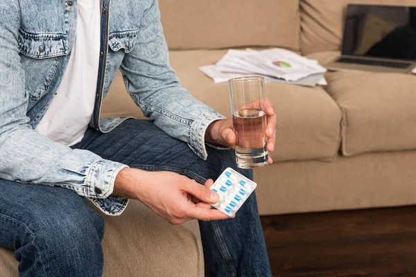 Cropped view of man holding blister with pills and glass of water near laptop and papers on couch