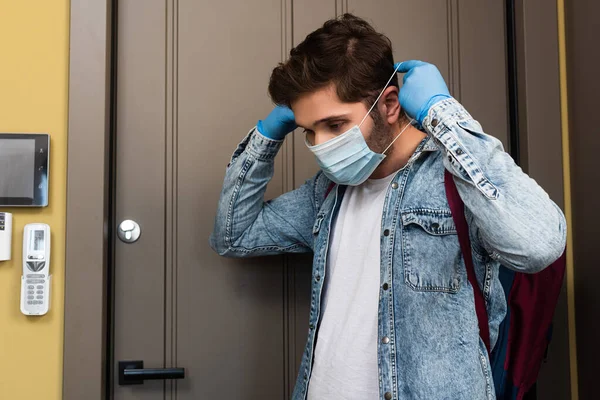 身穿乳胶手套的年轻人在走廊门边戴上医疗面罩 — 图库照片