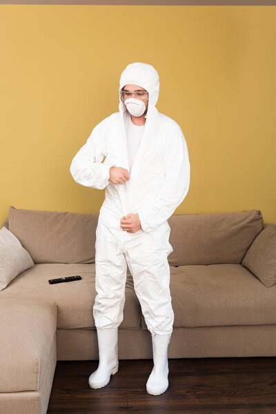 Young man in safety goggles and medical mask wearing hazmat suit at home 