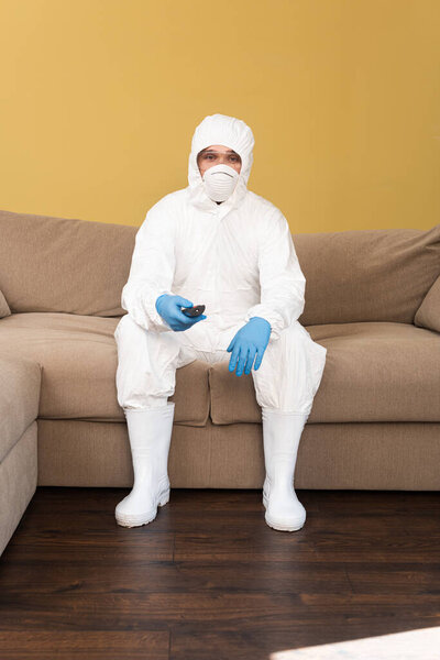 Man in hazmat suit, latex gloves and medical mask holding remote controller on sofa 
