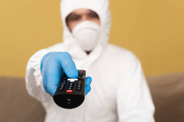 Selective focus of man in latex glove, hazmat suit and medical mask holding remote controller at home 