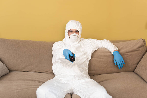 Man in medical mask, safety goggles and hazmat suit holding remote controller on sofa 
