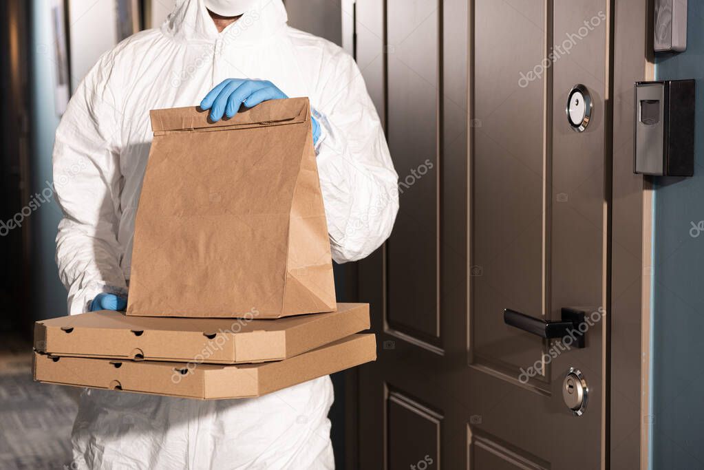 Cropped view of courier in hazmat suit and latex gloves holding package and pizza boxes near door on porch 