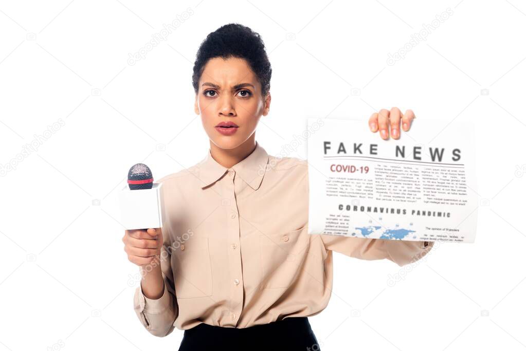 worried african american journalist with microphone showing newspaper with fake news covid-19 text isolated on white