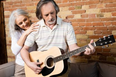 Sakallı ve olgun bir adam gülümseyen karısının yanında akustik gitar çalıyor. 