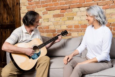 Sakallı ve olgun bir adam mutlu bir eşin yanında akustik gitar çalıyor. 