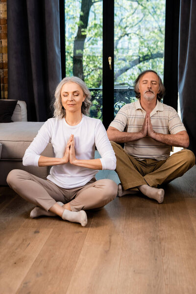 relaxed mature couple with closed eyes and praying hands sitting in lotus pose 