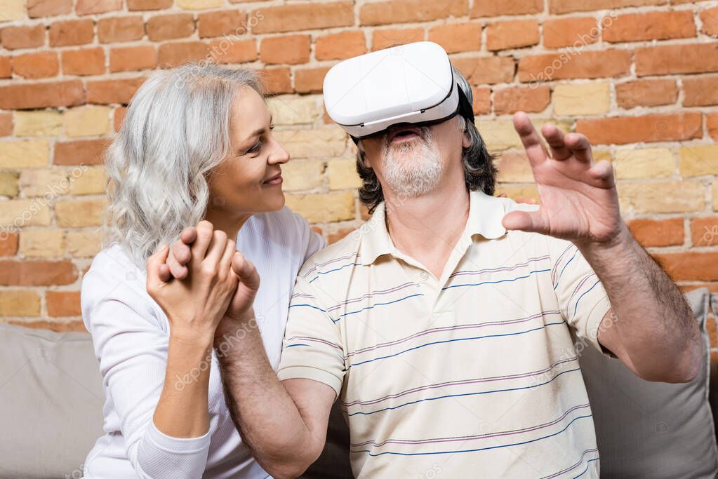 happy and mature woman looking at husband in virtual reality headset gesturing at home 