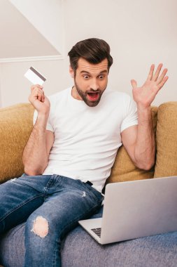 Kanepede kredi kartı tutarken dizüstü bilgisayara bakan şok olmuş bir adam.