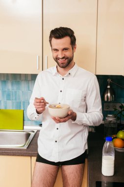 Gömlekli ve külotlu yakışıklı adam mutfakta mısır gevreği yerken kameraya gülümsüyor. 