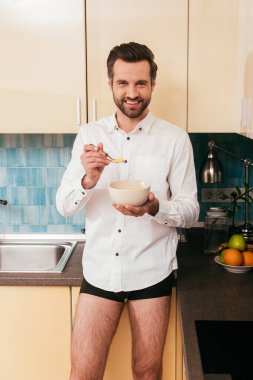 Gömlekli ve külotlu bir adam mutfakta mısır gevreği tutuyor. 
