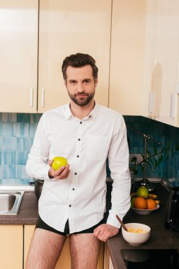 Külotlu ve gömlekli yakışıklı adam mutfak atölyesinde mısır gevreğinin yanında elma tutuyor. 