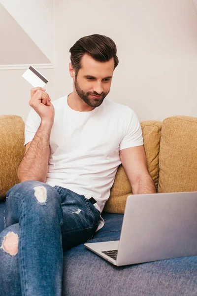 英俊的男人拿着信用卡在沙发上使用笔记本电脑 — 图库照片