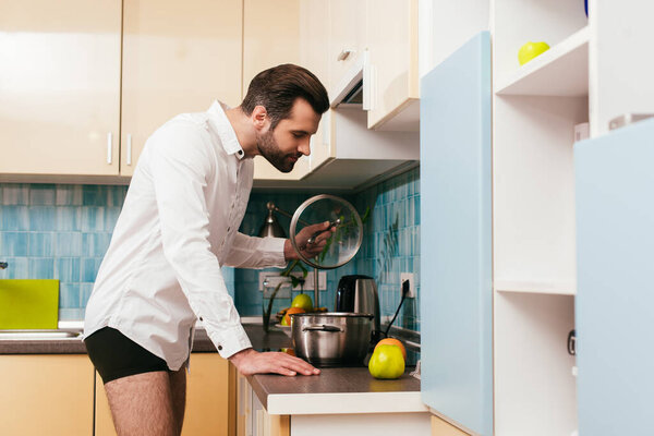 Side view of man in panties and shirt holding pan lid while cooking in kitchen 