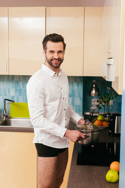 Side view of handsome man in shirt and panties holding pan and smiling at camera in kitchen 