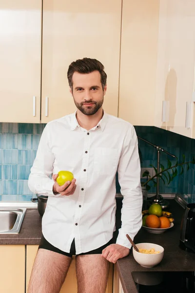 ハンサムな男でパンティーとシャツを保持していますリンゴシリアル近くキッチンワークトップ — ストック写真