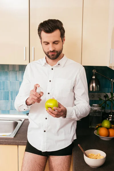 Man in shirt and panties holding apple and hand sanitizer near cereals in kitchen