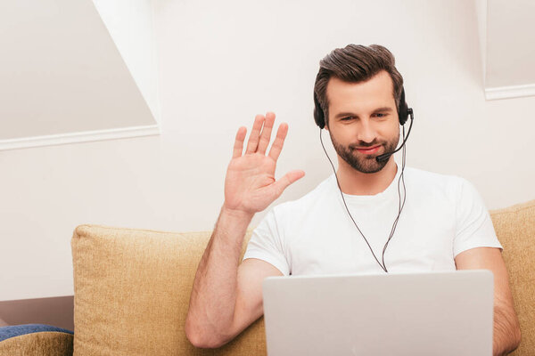 Selective focus of smiling teleworker in headset waving hand during video chat on laptop at home 