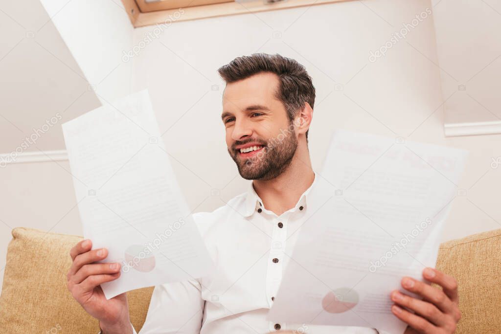 Selective focus of cheerful freelancer holding papers on sofa at home 