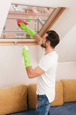 Oturma odasındaki pencereyi temizlerken deterjan ve bez kullanan adamın yan görüntüsü