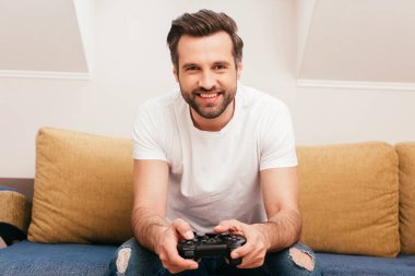 KYIV, UKRAINE - 14 Nisan 2020: Kameraya gülümseyen ve evdeki kanepede joystick tutan yakışıklı adam 