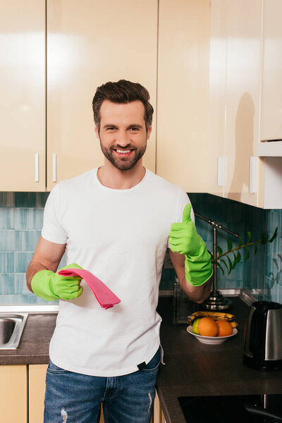 Smiling man in rubber gloves holding rad and showing like gesture in kitchen 