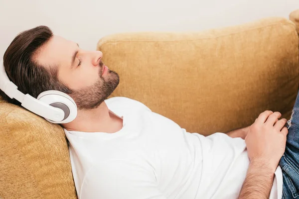 ソファに横たわっている間にヘッドフォンで音楽を聴くハンサムな男の側面図 — ストック写真