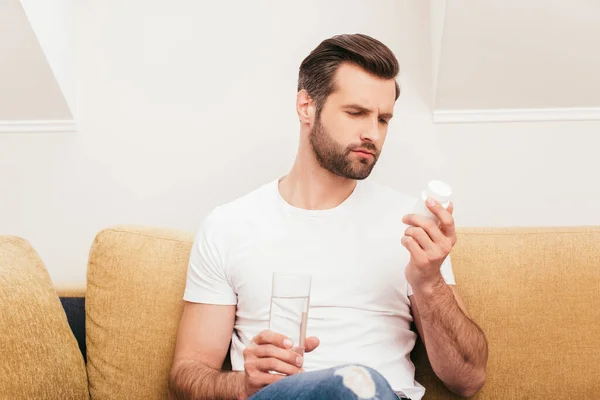 英俊男子拿着装有药丸和杯子的罐子躺在沙发上的选择性焦点 — 图库照片