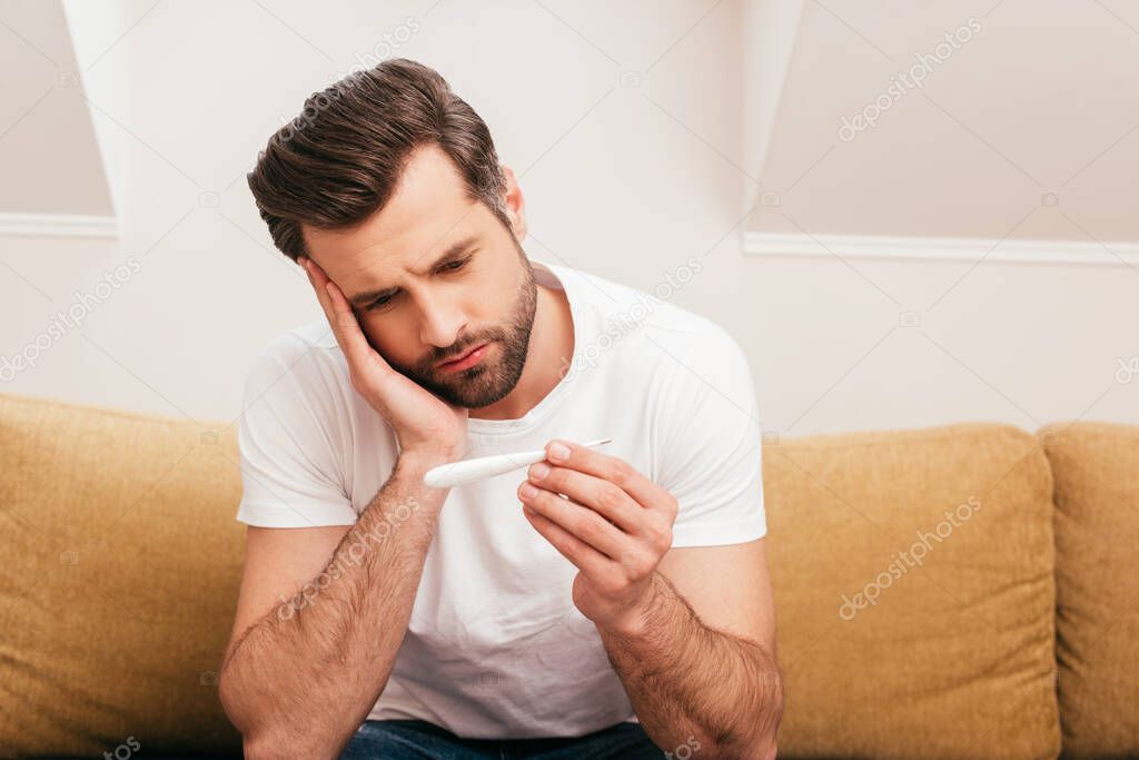 Upset man holding thermometer on sofa at home 