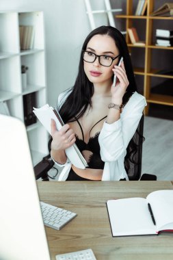 Sütyen ve tişörtlü seksi sekreterin seçici odak noktası. Akıllı telefonla konuşuyor ve çalışma masasında belgeleri tutuyor. 
