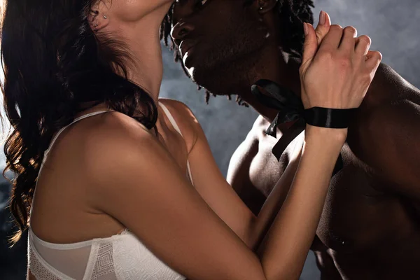 cropped view of seductive african american man hugging and going to kiss woman with bound hands on bed in dark with smoke